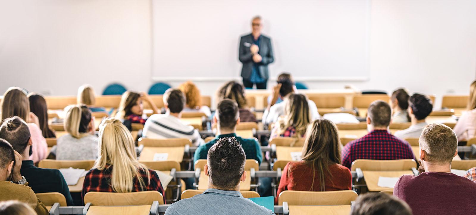 A classroom filled with students seated and facing a lecturer at the front. The lecturer is standing and speaking, with students attentively listening. The focus is on the back of the students' heads, highlighting their engagement in the lesson.